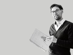 Schwarz-weiß-Porträt: Eine Person mit Brille und Mütze steht halber Oberkörper nach rechts im Bild und hält ein großes Buch mit Illustration auf dem Einband. Viel leere, hellgraue Fläche links