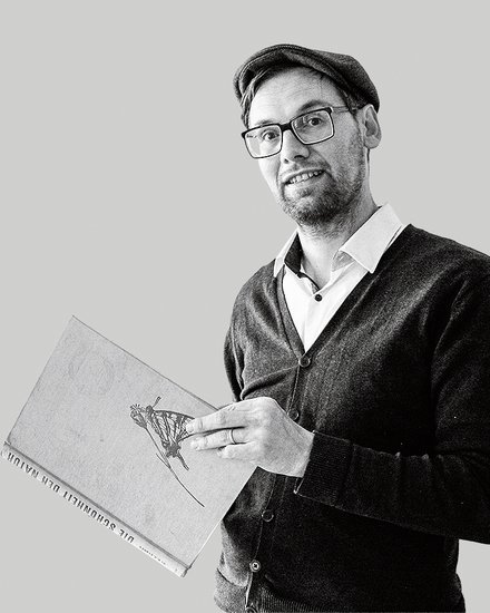 Schwarz-weiß-Porträt: Eine Person mit Brille und Mütze steht halber Oberkörper nach rechts im Bild und hält ein großes Buch mit Illustration auf dem Einband. Viel leere, hellgraue Fläche links