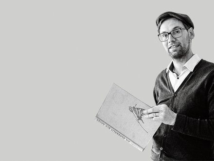 Schwarz-weiß-Porträt: Eine Person mit Brille und Mütze steht halber Oberkörper nach rechts im Bild und hält ein großes Buch mit Illustration auf dem Einband. Viel leere, hellgraue Fläche links