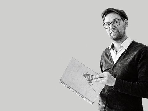 Schwarz-weiß-Porträt: Eine Person mit Brille und Mütze steht halber Oberkörper nach rechts im Bild und hält ein großes Buch mit Illustration auf dem Einband. Viel leere, hellgraue Fläche links
