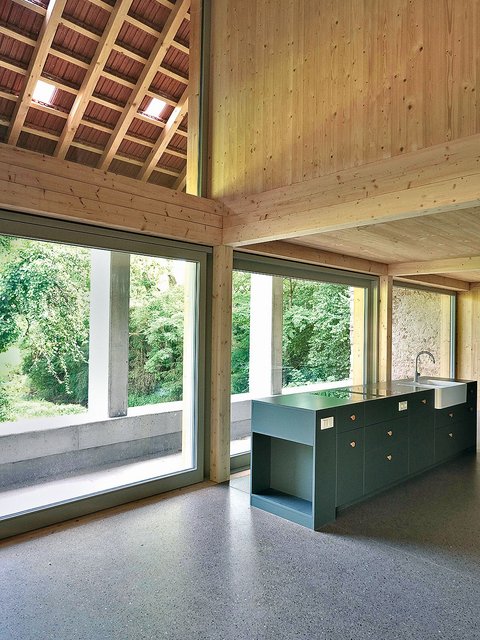 Innenraum mit großer Fensterfront, Holzwänden und -decke sowie einer Kücheninsel mit Spüle