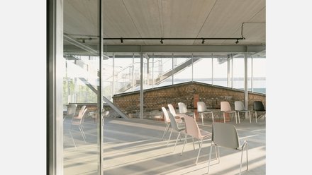 Heller Raum mit Glaswänden und Sichtbetondecke; lose gestellte Stühle im Halbkreis vor niedriger Backsteinmauer, Außentreppe durch Fenster sichtbar.