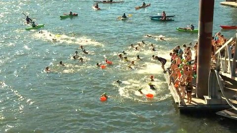 Viele Menschen schwimmen an einem Flussufer, während weitere Personen von einem Steg aus ins Wasser gehen. Kajaks begleiten das Geschehen auf dem Wasser.