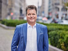 Porträt eines Mannes im blauen Anzug und weißen Hemd auf einer begrünten Stadtstraße mit unscharfem Hintergrund