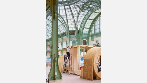 "Innenansicht einer klassischen Jugendstil-Ausstellungshalle mit grünen Stahlträgern und großem Glaskuppeldach. Im Raum stehen Holzgerüste und Konstruktionen im Bau. Ein Mensch auf einer Leiter arbeitet an einer Struktur."