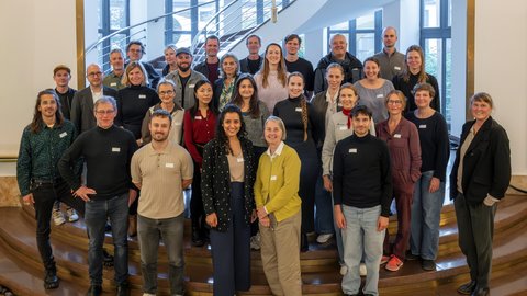 Ein Gruppenfoto von etwa 30 Personen unterschiedlichen Alters in einem hellen, großzügigen Foyer. Die Gruppe ist auf und vor den untersten Stufen einer elegant geschwungenen Treppe positioniert. Alle blicken lächelnd in die Kamera. Die Kleidung ist sportlich-elegant. Im Hintergrund ist die Architektur des Treppenaufgangs mit Metallgeländer und hohen Fenstern zu sehen.