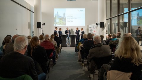: "Panel-Diskussion mit fünf Personen am Tisch. Im Hintergrund großer Beamer-Bildschirm mit Projektinformationen. Publikum sitzt im Vordergrund, einige Personen tragen Jacken."