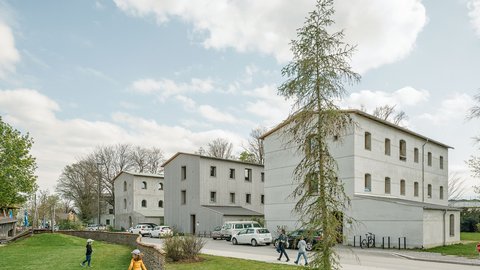 nsicht von drei minimalistischen, grau verputzten Mehrfamilienhäusern mit Satteldach. Im Vordergrund spielen Kinder auf einer Rasenfläche, im Hintergrund stehen einige Autos.