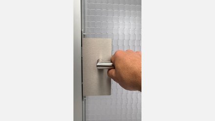 Hand greift einen rechteckigen, gebürsteten Metall-Türgriff vor einer weiß-gemusterten Wand