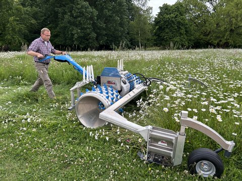  Ein Mann in einem karierten Hemd und beigefarbenen Hosen bedient ein spezielles, ferngesteuertes Mähgerät der Marke "Biber" auf einer Wiese mit Gänseblümchen. Die Maschine hat eine große blaue Walze mit spitzen Zacken und eine Metalleinheit. Der Hintergrund ist ein Waldrand.