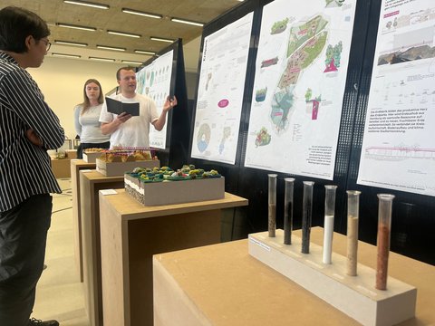 Ein Mann präsentiert vor zwei Zuhörerinnen einen Entwurf für einen „Erdpark“. Neben großformatigen Plänen stehen Landschaftsmodelle und mit verschiedenen Bodenproben gefüllte Glasröhren.