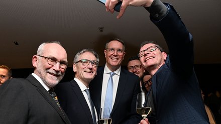 Gruppe von Teilnehmenden im Anzug macht ein Selfie; alle lächeln, zwei halten Weißweingläser.