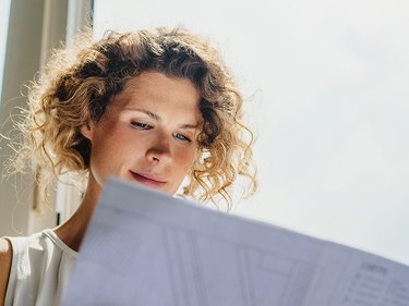 Person mit lockigem Haar hält ein großes Blatt mit technischen Zeichnungen oder Plänen vor sich.
