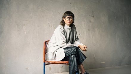 Sitzporträt einer erwachsenen Person mit Brille auf einem Stuhl; helles, weites Oberteil und dunkle Hose vor strukturierter grauer Wand.