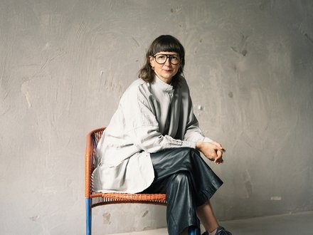 Sitzporträt einer erwachsenen Person mit Brille auf einem Stuhl; helles, weites Oberteil und dunkle Hose vor strukturierter grauer Wand.