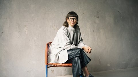 Sitzporträt einer erwachsenen Person mit Brille auf einem Stuhl; helles, weites Oberteil und dunkle Hose vor strukturierter grauer Wand.