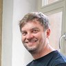 "Eine Frau mit schulterlangem blondem Haar und ein Mann mit braunem Haar stehen lächelnd in einem rustikalen Büro. Die Frau trägt ein rot-weiß kariertes Tweed-Sakko über einem dunklen Oberteil. Der Mann trägt ein schwarzes T-Shirt. Hinter ihnen ist eine Wand mit verputzter, abgebröckelter Oberfläche in Beige und Blaugrün sichtbar. An dieser Wand hängen mehrere architektonische Pläne und Renderings von Gebäuden. Auf dem Tisch vor ihnen liegen Baupläne, Modelle und farbige Entwurfsskizzen. Links steht eine beige Modellbox, rechts sind weitere Planunterlagen zu sehen. Das Fenster auf der rechten Seite lässt natürliches Licht in den Raum fallen."