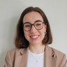 "Lächelnde Frau mit schulterlangen braunen Haaren und Brille. Sie trägt einen beigen Blazer über einem hellblauen Hemd und hat ihre Arme verschränkt. Der Hintergrund ist neutral grau."