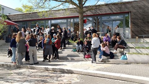 Gruppen von Menschen stehen und sitzen in der Sonne auf breiten Außentreppen vor einem Campusgebäude; Pausengespräch unter Bäumen.