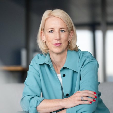 Hellblonde Frau in blau‑türkiser Bluse sitzt mit verschränkten Armen auf den Knien, blickt direkt in die Kamera; dezentes Make‑up, rote Fingernägel, Uhr und Ring sichtbar, unscharfer Innenraumhintergrund.