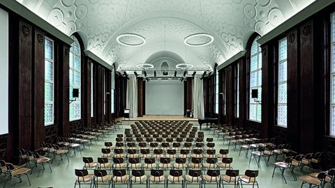 Großer Saal mit hohen Fenstern, gewölbter Decke und Reihen von Stühlen vor einer Bühne mit Klavier
