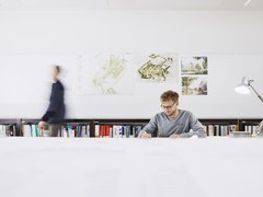 Mann sitzt an langem Schreibtisch mit Büchern im Hintergrund und einer Schreibtischlampe rechts neben ihm. 