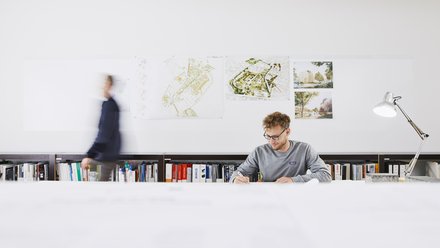 Mann sitzt an langem Schreibtisch mit Büchern im Hintergrund und einer Schreibtischlampe rechts neben ihm. 