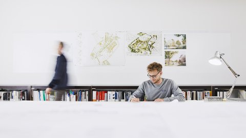 Mann sitzt an langem Schreibtisch mit Büchern im Hintergrund und einer Schreibtischlampe rechts neben ihm. 