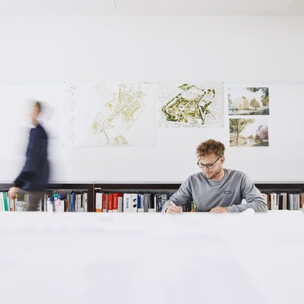 Mann sitzt an langem Schreibtisch mit Büchern im Hintergrund und einer Schreibtischlampe rechts neben ihm. 
