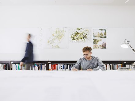 Mann sitzt an langem Schreibtisch mit Büchern im Hintergrund und einer Schreibtischlampe rechts neben ihm. 