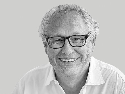 Schwarzweiß-Nahaufnahme: lächelnder Mann mit Brille und weißem Hemd vor neutral grauem Hintergrund.