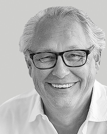 Schwarzweiß-Nahaufnahme: lächelnder Mann mit Brille und weißem Hemd vor neutral grauem Hintergrund.