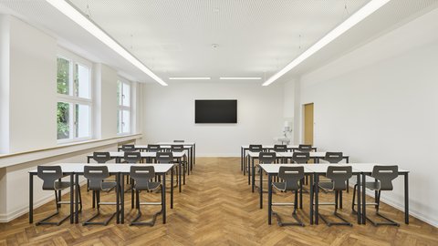 Leerer Klassenraum mit Holzparkett, mehreren Reihen schwarzer Stühle und Tische, großen Fenstern links und einem Bildschirm an der weißen Wand vorne