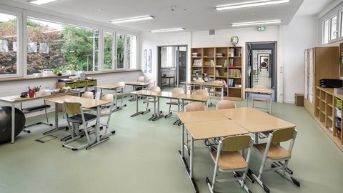 Heller Klassenraum mit mehreren Tischen und Stühlen, großen Fenstern und Regalen an den Wänden