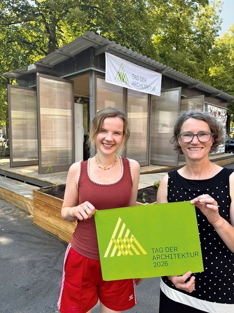 „Zwei lächelnde Frauen halten ein grünes Schild mit der Aufschrift ‚Tag der Architektur 2026‘ vor dem fertigen Pavillon mit transluzenten Flügeltüren.“