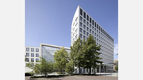 "Modernes Gebäudeensemble mit Wohn- und Geschäftshäusern. Weißes und graubetontes Hochhaus rechts mit regelmäßigen Fensterreihen. Niedrigeres weißes Gebäude mit großflächig verglaster Fassade links. Grüne Bäume im Vordergrund, gepflasterter öffentlicher Platz. Blauer Himmel."