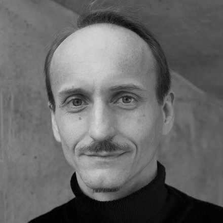 Schwarz‑weiß‑Porträt eines erwachsenen Mannes im Rollkragen vor Betonwand; neutraler Blick.