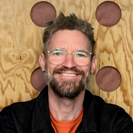 Porträt eines lächelnden Mannes mit Brille vor einer Holzplatte mit runden Einlagen.