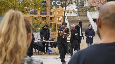 "Ein Rapper in schwarzem Trikot und Kappe tritt im Freien auf und singt in ein Mikrofon. Im Hintergrund bedient ein DJ ein Mischpult, dahinter steht ein hohes Holzgerüst. Im Vordergrund sieht man unscharf die Köpfe des Publikums."