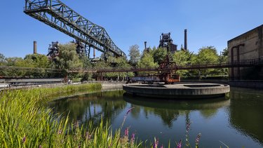 Eine farbige Aufnahme zeigt eine ehemalige Industrieanlage mit Wasserbecken, Rohrleitungen, Förderbrücken und Hochöfen, die heute von Vegetation umgeben sind, während Wege und Stege den Raum erschließen, wodurch das Bild den Wandel von industrieller Produktionsstätte zu einem öffentlich zugänglichen Landschafts- und Kulturraum sichtbar macht und für Transformation, Nachnutzung und die Zukunftsfähigkeit von Industriebauten steht.
