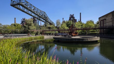 Eine farbige Aufnahme zeigt eine ehemalige Industrieanlage mit Wasserbecken, Rohrleitungen, Förderbrücken und Hochöfen, die heute von Vegetation umgeben sind, während Wege und Stege den Raum erschließen, wodurch das Bild den Wandel von industrieller Produktionsstätte zu einem öffentlich zugänglichen Landschafts- und Kulturraum sichtbar macht und für Transformation, Nachnutzung und die Zukunftsfähigkeit von Industriebauten steht.