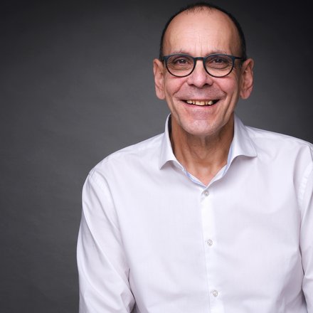 Porträt eines lächelnden Mannes mit Brille und weißem Hemd vor dunklem Hintergrund.