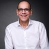 Porträt eines lächelnden Mannes mit Brille und weißem Hemd vor dunklem Hintergrund.
