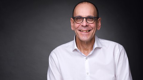 Porträt eines lächelnden Mannes mit Brille und weißem Hemd vor dunklem Hintergrund.
