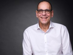 Porträt eines lächelnden Mannes mit Brille und weißem Hemd vor dunklem Hintergrund.