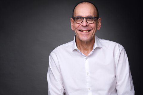Porträt eines lächelnden Mannes mit Brille und weißem Hemd vor dunklem Hintergrund.
