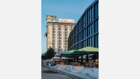 Blick auf ein rundes Hochhaus mit mehreren Fensterreihen und einem großen Kellogg’s-Logo auf dem Dach; rechts davon ein modernes Glasgebäude mit blauer Fensterfront. Im Vordergrund eine Außenterrasse mit grünen Sonnenschirmen, Holztischen und niedrigen Holzsitzstufen, klarer blauer Himmel.