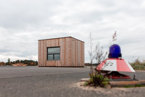 Kleines Holzgebäude auf freier Fläche, im Vordergrund ein rotes Baustellenlicht.