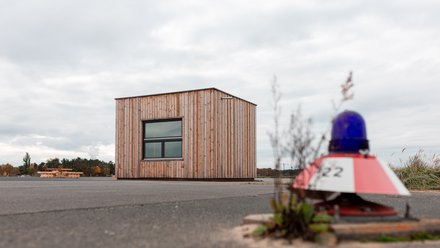 Kleines Holzgebäude auf freier Fläche, im Vordergrund ein rotes Baustellenlicht.
