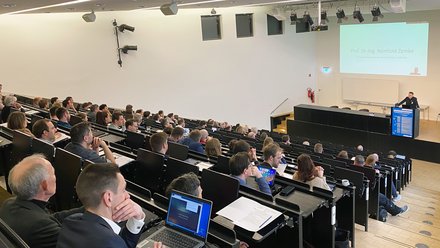 Großer Hörsaal mit ansteigenden Sitzreihen, in denen zahlreiche Teilnehmende sitzen. Vorn steht ein Redner am Pult, hinter ihm ist eine Präsentationsfolie auf eine Leinwand projiziert.